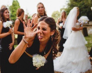 A woman playfully makes a funny face towards the camera, holding a bouquet of white flowers. Behind her, bridesmaids in black dresses and a bride in a white gown are standing outdoors—a perfect candid moment captured by talented Somerset wedding photographers.