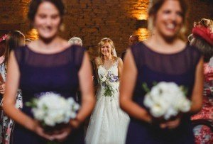 A bride holding a bouquet smiles, walking down an aisle. In the foreground, two bridesmaids in dark dresses, also holding bouquets, are slightly blurred. The background shows a warmly lit stone wall—a perfect capture by documentary wedding photographers capturing every cherished moment.