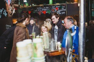 People are gathered at a market stall, hot drinks in hand. Cup stacks line the counter while a chalkboard menu sets the scene. Its cold, but the bustling crowd and cozy vibes would charm even wedding photographers from Somerset seeking inspiration in authentic moments.