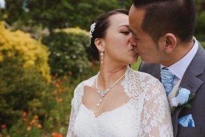A bride and groom share a kiss outdoors, captured beautifully by Bristol wedding photographers. She is in a white lace dress and jewelry, while he wears a gray suit with a light blue tie and flower boutonniere. They stand surrounded by lush greenery and vibrant orange flowers.