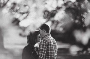 Black and white image of a couple standing closely, facing each other outdoors, captured by wedding photographers in Bristol. The background is softly blurred, creating a dreamy effect. The man is wearing a plaid shirt, and the woman has glasses and long hair. Both are smiling gently.