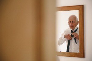 A man in a white shirt is tying a black tie while looking in a wooden-framed mirror, reminiscent of the candid style favored by documentary wedding photographers. The reflection shows him concentrating, with the foreground slightly blurred to emphasize the mirrors image.