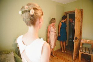 A bride with flowers in her updo stands in a room, facing two women and a man at the door. The woman in pink and the woman in blue smile, while the man leans in with a cheerful expression. Captured by talented documentary wedding photographers, the scene unfolds against light green walls and wooden floors.