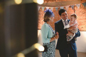 A man in a suit happily holds a smiling baby. A woman in a floral dress with a blue cardigan and fascinator stands beside them, captured beautifully by documentary wedding photographers. They are indoors, with brick walls and string lights in the foreground.