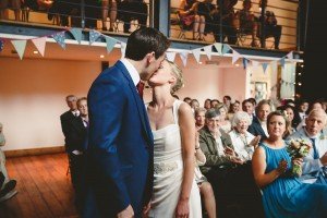 A bride and groom share a kiss in front of seated guests during an indoor ceremony captured beautifully by documentary wedding photographers. The groom is in a blue suit and the bride in a white gown, as bunting hangs overhead, with guests watching from both the ground and a balcony above.