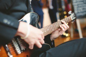 A person wearing a black suit plays an electric guitar with focus on the hands and guitar, capturing a scene reminiscent of documentary wedding photographers. The background is softly blurred with hints of lights and people, creating a warm, ambient atmosphere.