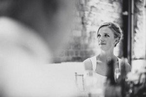 Black and white photo of a woman in a wedding dress smiling and looking to the side. Captured by one of the finest Bristol wedding photographers, she sits indoors with blurred details in the foreground, creating a soft, intimate atmosphere.
