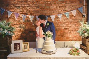 A couple is kissing in front of a brick wall decorated with triangular bunting. Behind their white tiered wedding cake adorned with flowers, they relish the moment captured by documentary wedding photographers. Framed photos and flower arrangements elegantly grace the table.