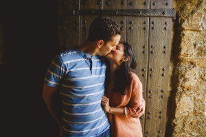 A couple kissing in front of a rustic, wooden door, captured beautifully by Somerset wedding photographers. The man sports a blue striped shirt, and the woman wears a pink sweater. Theyre in a stone building, with light softly illuminating the scene.