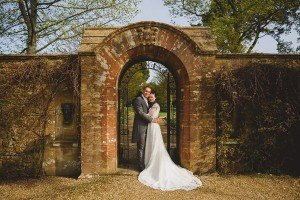 A bride and groom stand embracing under a rustic stone archway, captured artfully by wedding photographers Somerset trusts. The bride wears a long white gown, and the groom is dressed in a dark suit. The background features leafy trees and weathered stonework.