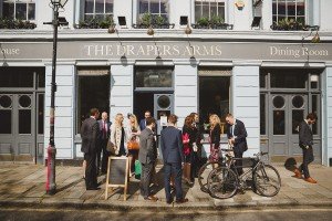 A group of well-dressed people stands outside The Drapers Arms pub, engaging in conversation. A bicycle is parked nearby, and the building features large windows and double doors—an ideal scene for wedding photographers in the Cotswolds to capture a moment of candid elegance.
