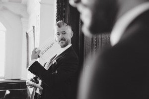 Black and white image of a man in a suit holding a HOORAY sign. He has a beard and gazes at the camera, while another person is blurred in the foreground. Captured by Bristol wedding photographers, they stand in an elegant room with ornate details.