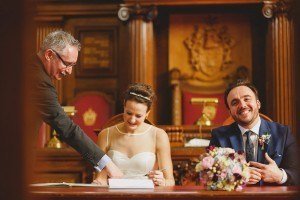 In a grand room adorned with wooden decor and a coat of arms, the bride in her white dress signs a document, laughing. The groom in his suit smiles as he watches. A man in glasses leans over to point at the paper, capturing a moment that wedding photographers Somerset would adore, while a bouquet sits nearby.