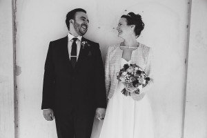 A smiling couple faces each other, holding hands. The man wears a dark suit and tie with a boutonniere, while the woman in a light-colored dress holds a bouquet. Captured by documentary wedding photographers, the backdrop is a simple, light-colored wall, and the mood is joyfully serene.