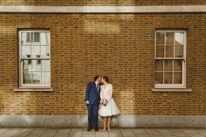 A couple in formal attire kisses in front of a brick wall with two windows. The woman wears a white dress, carrying a bouquet, while the man dons a blue suit. Illuminated by sunlight on the paved sidewalk, they capture a timeless moment perfect for any documentary wedding photographers portfolio.