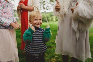 A young child with blonde hair, in a striped sweater, gives a thumbs-up while standing between two adults doing the same. Theyre outdoors amidst green grass and trees, perfectly capturing the joyful vibe typical of a Bristol wedding photographers work.