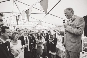 A grayscale image captures a lively wedding reception, artfully documented by a talented Cotswolds wedding photographer. A man gives a speech with a microphone, smiling amid guests who attentively listen and smile. The venue is elegantly adorned with hanging flags and lights.