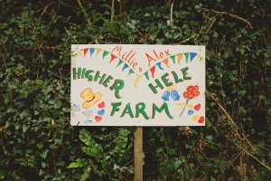 A colorful wedding sign reads Millie & Alex, Higher Hele Farm surrounded by hand-drawn hearts, flowers, a top hat, and flutes, with greenery in the background. Captured beautifully by Somerset wedding photographers, it perfectly sets the tone for this enchanting celebration.