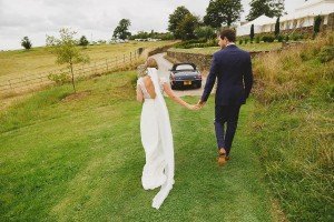 A bride and groom walk hand in hand on a grassy path, captured beautifully by Somerset wedding photographers. The bride wears a white gown with lace details, and the groom is in a dark suit. Ahead, a convertible car is parked, framed by trees and a building in the background.