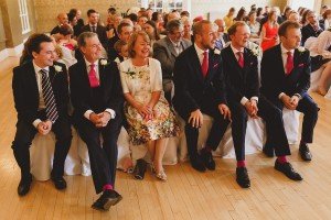 At a formal indoor event, a group sits in a row. The men don bright pink ties and matching socks, while one person sports a floral dress. In the background, the crowd is seated attentively. This elegant scene captures the attention of Somerset wedding photographers with its vivid detail.