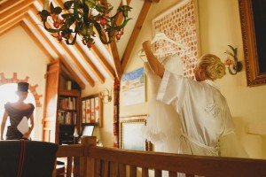 A woman in a white robe labeled Bride holds a wedding dress on a hanger in a warmly lit room with wooden beams, captured beautifully by Somerset wedding photographers. Another person in a hat stands nearby, surrounded by a chandelier, bookshelf, and framed art that enrich the space.
