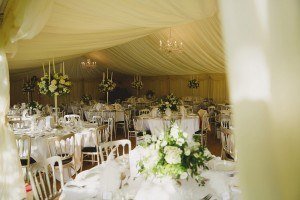 The interior of a wedding tent features elegant white drapery. Tables are adorned with white tablecloths, floral centerpieces, and surrounded by white chairs. Chandeliers hang from the ceiling, casting a warm glow, perfectly capturing the magic for documentary wedding photographers in Bristol.