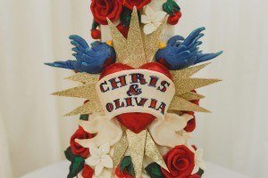 A wedding cake decoration featuring a red heart with Chris & Olivia in black letters, surrounded by gold glitter stars, red roses, white flowers, and two blue birds. Perfect for photos by Bristol wedding photographers capturing the charm of your special day.