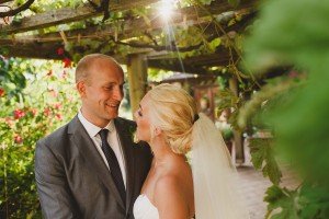 A bride and groom stand closely under a vine-covered pergola as sunlight filters through the leaves, creating a romantic atmosphere. Captured by Somerset wedding photographers, the groom in a gray suit smiles warmly at his bride, who is radiant in her white dress and veil.