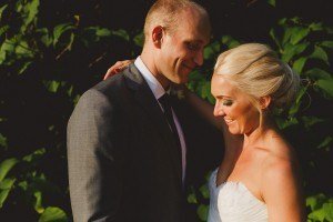 Bride and groom smiling warmly at each other, surrounded by lush green foliage. The bride has blonde hair and is wearing a white strapless dress. The groom is in a gray suit. Captured by Somerset wedding photographers, the lighting creates a soft and intimate atmosphere.