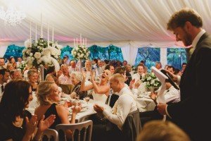 A joyful wedding reception inside a decorated tent captures the lens of Somersets finest wedding photographers. Guests are seated around tables with floral centerpieces, clapping and laughing, as a man gives a speech in the foreground. The atmosphere is festive and lively.