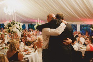 Two people embrace in a warmly lit wedding reception under a large tent expertly captured by a Bristol wedding photographer. Guests sit at round tables adorned with white flowers, watching the hug with smiles. The atmosphere is joyful and celebratory.