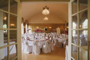 The elegant banquet hall, perfect for wedding photographers from Bristol or Somerset, boasts chandeliers and an arched ceiling. It’s set for an event with round tables draped in white cloths and chairs with white sashes. Tall floral centerpieces with red flowers and candelabras complete the scene.