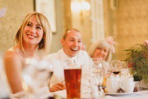 A smiling woman in a white dress sits at a table set with glasses and a beer. Beside her, a man in a white shirt grins, while a blurred guest lingers behind. Captured by accomplished documentary wedding photographers, the scene evokes the joy of such celebratory events.
