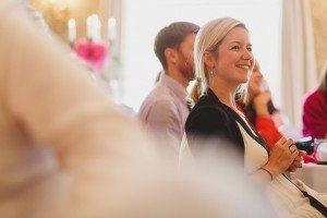 A woman smiling and sitting in a softly lit room, surrounded by blurred figures and decor. She holds a pen and notebook, as if attending a meeting or lecture. The ambiance resembles the warm, subtle style favored by wedding photographers in Bristol, creating an inviting backdrop.