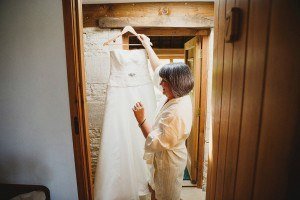 A woman in a robe admires a wedding dress hanging on a wooden hanger in a rustic, sunlit room with an open wooden door. Captured by Somerset wedding photographers, the strapless gown features a decorative belt, embodying timeless elegance.
