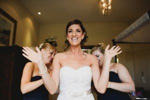 A joyful bride in a white strapless lace wedding dress stands with her arms raised, smiling widely. Two bridesmaids in dark dresses adjust her gown in the background. The room is softly lit with a chandelier overhead, captured beautifully by Somerset wedding photographers.