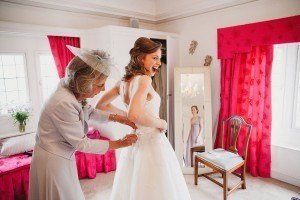 A bride in a white dress is gently assisted by an older woman in a light gray outfit and hat. The room, adorned with red curtains, features a mirror capturing the bride’s image. In the background, flowers are visible—a scene perfect for somerset wedding photographers to capture.