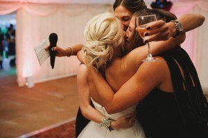 Three women in formal attire embrace tightly at an indoor event. One holds a microphone, another raises a wine glass, and smiles abound. Bristol wedding photographers capture the warm lighting and white draped backdrop that enhance the celebratory atmosphere perfectly.