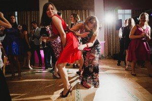 People are dancing energetically at a party. A woman in a red dress bends slightly forward while another in a floral dress playfully pulls her skirt. Other guests enjoy the lively atmosphere, creating perfect moments for documentary wedding photographers to capture amidst the vibrant celebration.