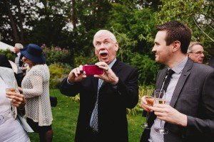 A man with white hair playfully takes a photo with a pink phone, surrounded by guests in formal attire holding drinks. They are outdoors, amidst lush greenery, capturing the candid spirit of the gathering—perfect for documentary wedding photographers at a charming garden party.