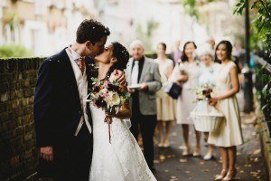 A bride and groom kiss on a leafy pathway, captured beautifully by Bristol wedding photographers. Surrounded by smiling guests, the bride holds a bouquet, while a girl with a flower basket stands nearby. The scene is joyful and elegant.