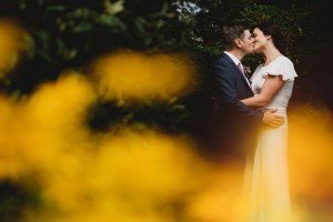 A couple stands closely, sharing a kiss outdoors. The person on the left is wearing a dark suit with a light shirt, while the person on the right is in a white dress. With yellow flowers blurred in the foreground, Somerset wedding photographers capture this romantic moment beautifully.