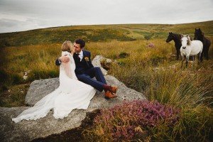 A bride and groom sit on a rock in a grassy field, embracing each other. The bride wears a white dress, and the groom is in a dark suit. Three horses stand nearby, adding rustic charm to the serene landscape captured beautifully by Wedding Photographers Somerset.