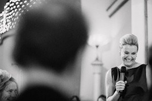 A black and white photo captures a woman smiling broadly while holding a microphone, speaking to an audience. The background is blurred, with lights softly glowing. A blurry figure in the foreground adds intimacy and joy, showcasing the talent of wedding photographers from Somerset at an event.