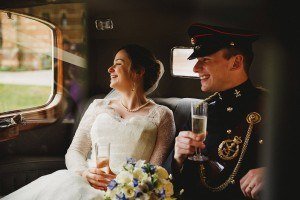 A bride in a white lace wedding dress and a groom in a formal military uniform smile and hold glasses of champagne inside a vintage car. Captured by Somerset wedding photographers, the bride has a bouquet of blue and white flowers on her lap as they both look out the window joyfully.