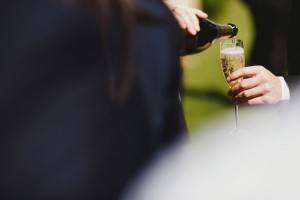 A person is pouring champagne into a glass outdoors, perfectly capturing the candid essence that documentary wedding photographers adore. The scene is partially out of focus, with one hand holding the glass and the other pouring from the bottle amid a blurry green garden backdrop.