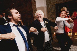 Three people in suits and a woman in a dark jacket are joyfully dancing at a lively event captured by Bristol wedding photographers. The older woman is smiling widely, and the two men flanking her are energetically moving, creating a fun and celebratory atmosphere.