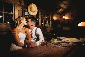 A couple in wedding attire shares a kiss while seated in a cozy, dimly lit wooden room. A pizza oven glows warmly in the background, and a cutting board with ingredients is on the table beside a mug of beer—perfectly captured by skilled documentary wedding photographers.