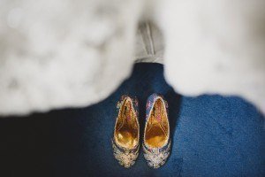 Ornate embroidered shoes with gold interiors rest on a deep blue carpet, partially framed by a billowing white fabric above—a scene perfectly captured by Somerset wedding photographers.