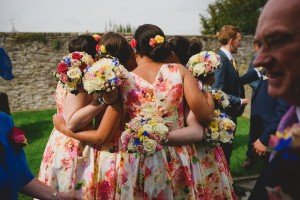 A group of people wearing floral dresses and holding colorful bouquets huddle together outdoors, captured artfully by a Somerset wedding photographer. The background features a stone wall and greenery, while onlookers, including a person in a suit, are visible nearby.